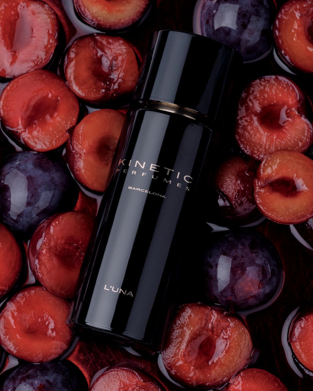 Kinetic Perfumes L'Una Extrait de Parfum 100 ml – schwarzer Flakon liegend auf halbierten roten Pflaumen