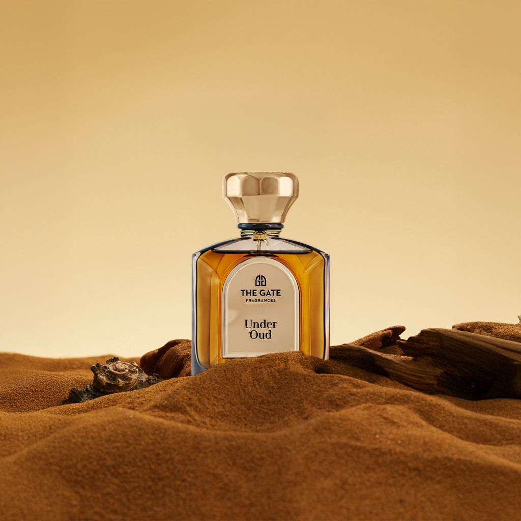 The Gate Under Oud Extrait de Parfum 100 ml – bernsteinbrauner Flakon auf Sanddüne mit Treibholz vor goldenem Hintergrund