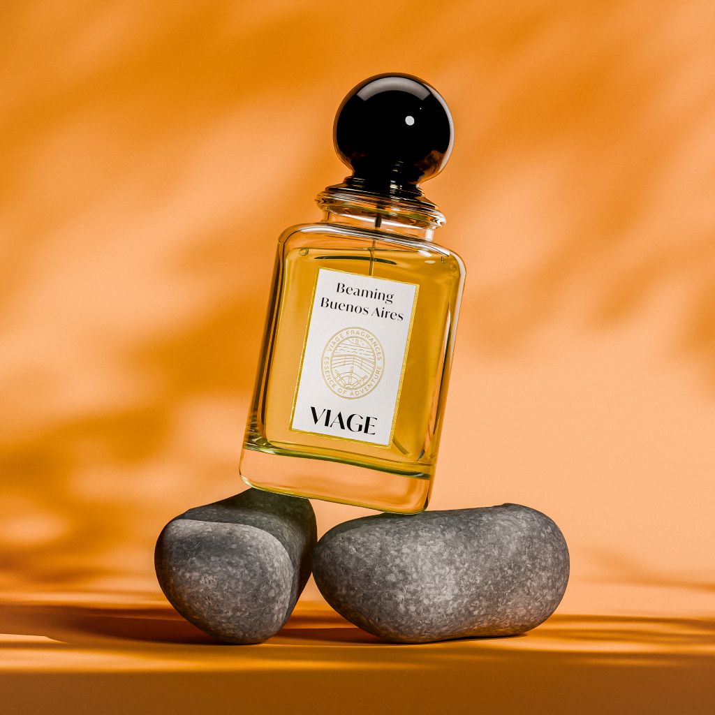 Viage Beaming Buenos Aires Extrait de Parfum 100 ml – Flakon schräg auf grauen Kieselsteinen vor orangem Hintergrund mit Blattschatten
